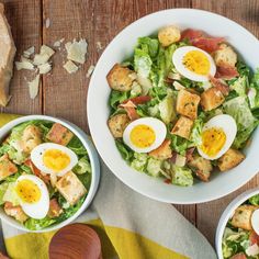 three bowls filled with salad and eggs on top of a wooden table next to bread