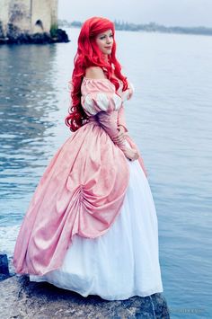 a woman in a pink and white dress standing on the edge of a body of water