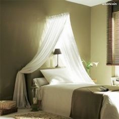 a bed with white sheets and pillows in a room next to a window covered by sheer curtains