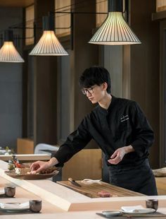 a man preparing food in a restaurant with lights hanging from the ceiling and wooden tables