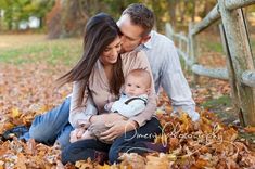 a man and woman holding a baby while sitting on leaves in front of a fence