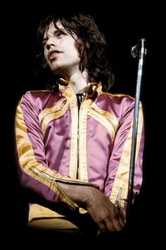 a man with long hair wearing a purple and gold jacket holding a microphone in his hand