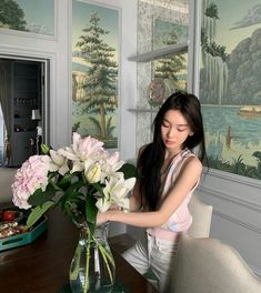 a woman arranging flowers in a vase on a dining room table next to a painting