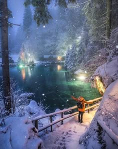 a person is standing on a bridge in the snow near a lake with lights and trees