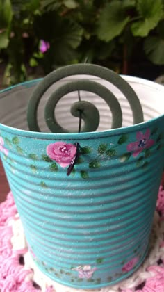 a blue tin can with pink flowers on it sitting on a doily next to a potted plant