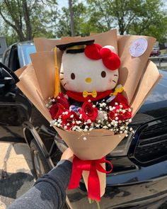 a hello kitty graduation bouquet being held by someone's hand in front of a car