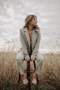 a woman sitting on top of a wooden stool in a field with tall grass behind her