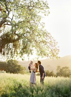 a man and woman holding a baby in a field under a tree with the sun behind them