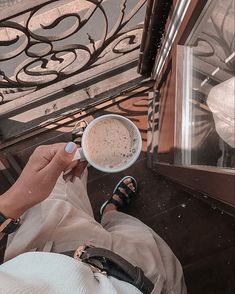 a person holding a cup of coffee in their hand while sitting on the ground outside