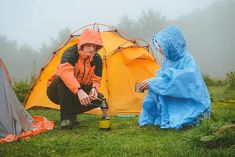 two people in raincoats are setting up their tents on the grass with one holding a cup