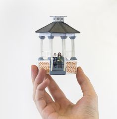 a hand holding up a miniature house with two people sitting on the porch in front of it