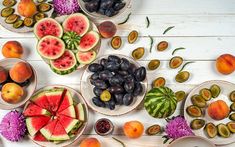 various fruits and vegetables are arranged in bowls on a white wooden surface, including watermelon, plums, grapes, peaches, and figs