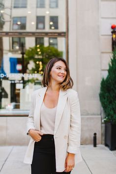 a woman in black pants and a white blazer is standing on the sidewalk smiling