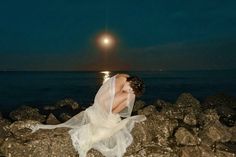 a woman in a white dress is sitting on rocks by the ocean at night with her head down