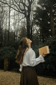 a woman holding a box in her hand and looking up into the sky with trees behind her