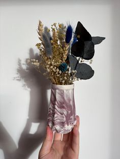 a hand holding a vase with flowers and feathers in it, against a white wall