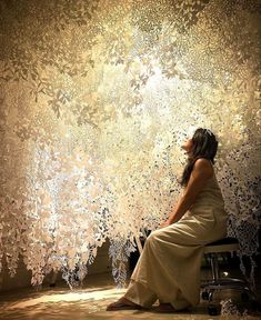 a woman sitting on a bench in front of a wall with intricate paper flowers hanging from it