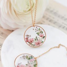 two necklaces with flowers on them sitting on top of a white table next to a flower