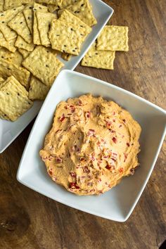 two white bowls filled with crackers and dip next to some chips on a wooden table