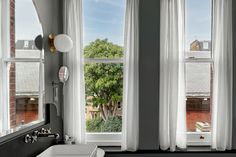 a bathroom with two windows and a sink in front of the window is white curtains