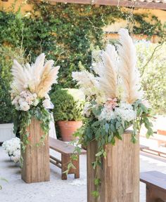 two tall vases with flowers and greenery on them are sitting in front of a bench