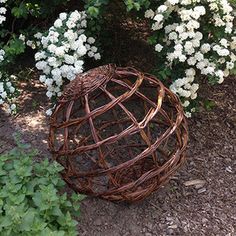 a round sculpture made out of wicker sitting in the middle of some bushes and flowers