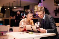 a man and woman sitting at a table with wine glasses looking at an open book