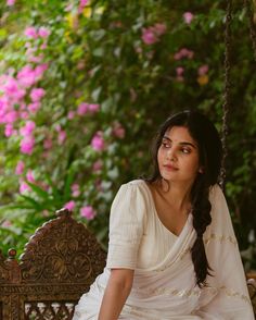 a woman sitting on a swing in front of pink flowers and greenery, wearing a white sari