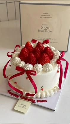 a white cake with strawberries on top and red ribbon around it, sitting next to a sign