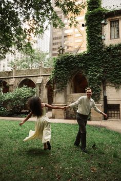 a man and woman holding hands while walking through the grass in front of an old building