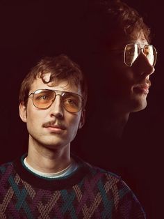 two men with glasses are looking at each other in front of a black background and one is wearing a sweater