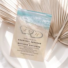 a wedding card with two hearts in the sand next to an open palm tree fan