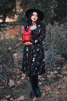 a woman wearing a black hat and dress standing in the woods with her hands on her chest