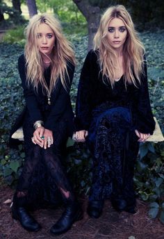 two women sitting on a bench next to each other in front of some bushes and trees