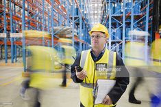 a man in a yellow safety vest and hard hat holding a clipboard while walking through a warehouse