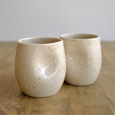 two white ceramic cups sitting on top of a wooden table next to each other and one has black speckles