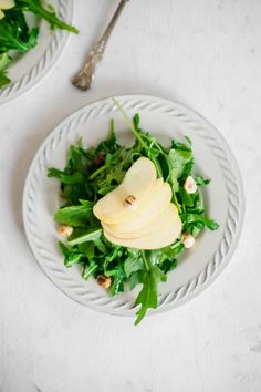 two white plates topped with green salad and sliced pears next to silverware on a white table