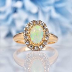 an opal and diamond ring sits on a white surface with blue flowers in the background