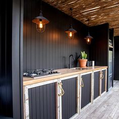 an outdoor kitchen with black walls and wooden flooring, hanging lights above the counter