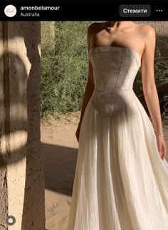 a woman in a white dress standing next to a tree and dirt area with trees behind her