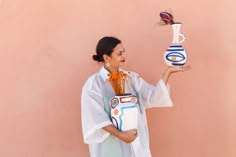 a woman is holding a vase with flowers in it while standing against a pink wall