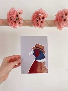 a hand holding a piece of paper with a painting of a bird on it and pink flowers in the background