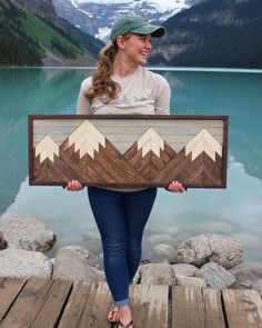 a woman holding up a wooden sign with mountains in the background and water below her