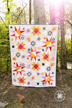 a colorful quilt hanging from the side of a tree in front of some trees and grass