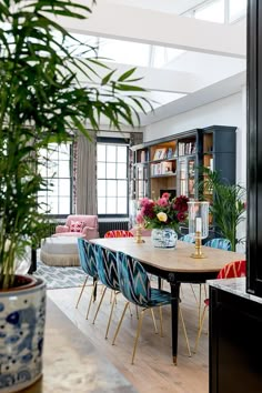 a dining room table surrounded by colorful chairs and potted plants in front of the bookshelf