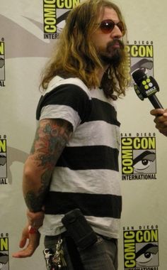 a man with long hair and tattoos holding a microphone in front of a sign that says comiccon