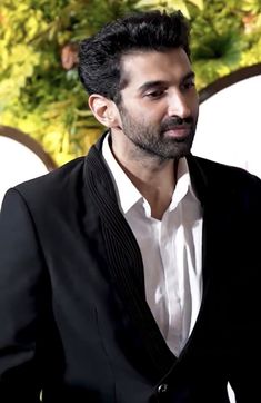 a man in a black suit and white shirt standing next to a wall with plants on it