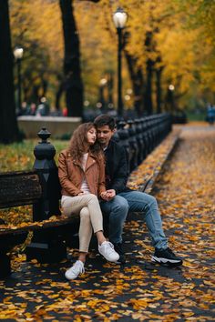 a man and woman are sitting on a bench in the park with leaves all around them