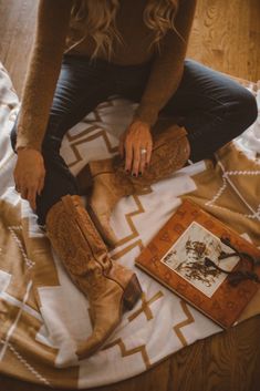 a woman sitting on top of a bed next to a book and some brown boots