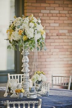 a table topped with a tall vase filled with white and yellow flowers on top of a blue table cloth